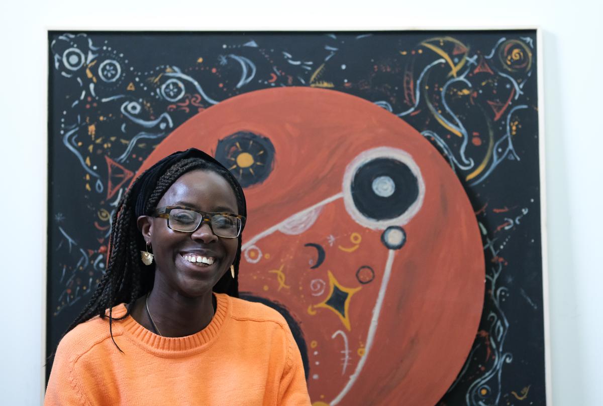 The artist standing in front of the painting, smiling at the camera, wearing an orange jumper.