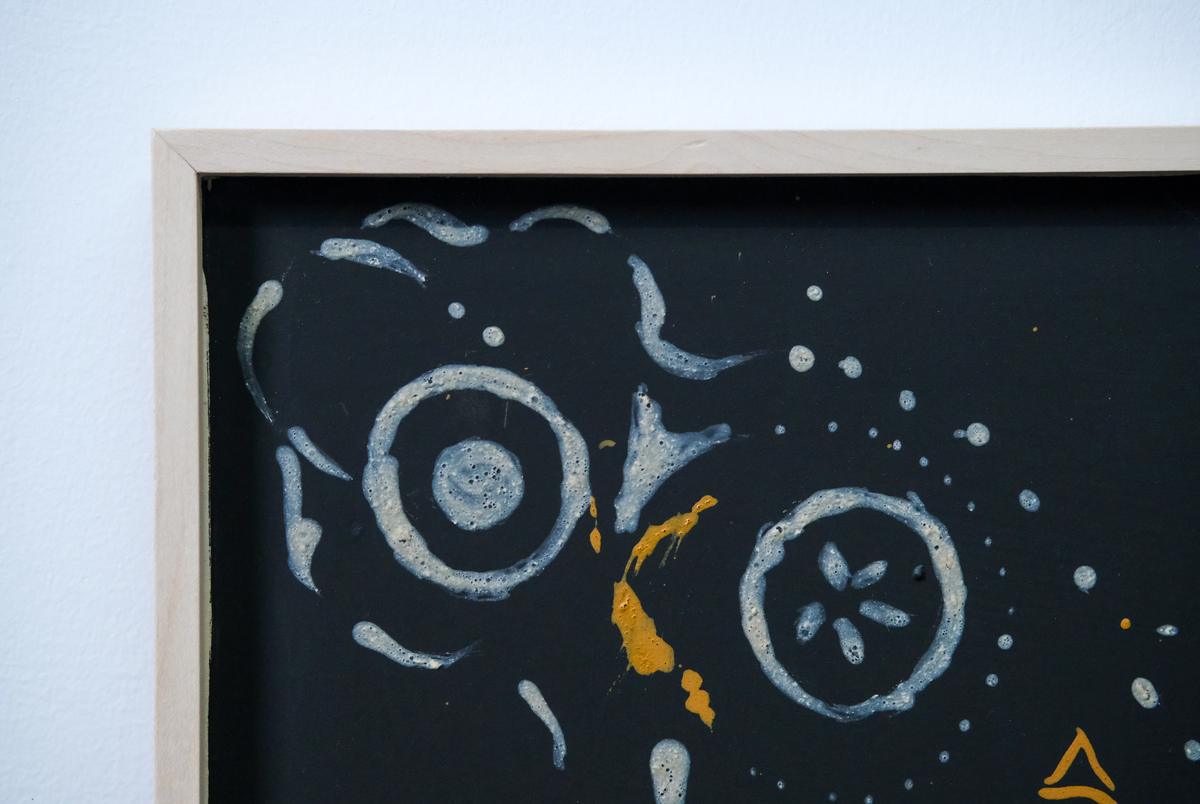 A close view of the framed corner: pale blue-grey rings and marks on the black ground, with a streak of yellow ochre between them.