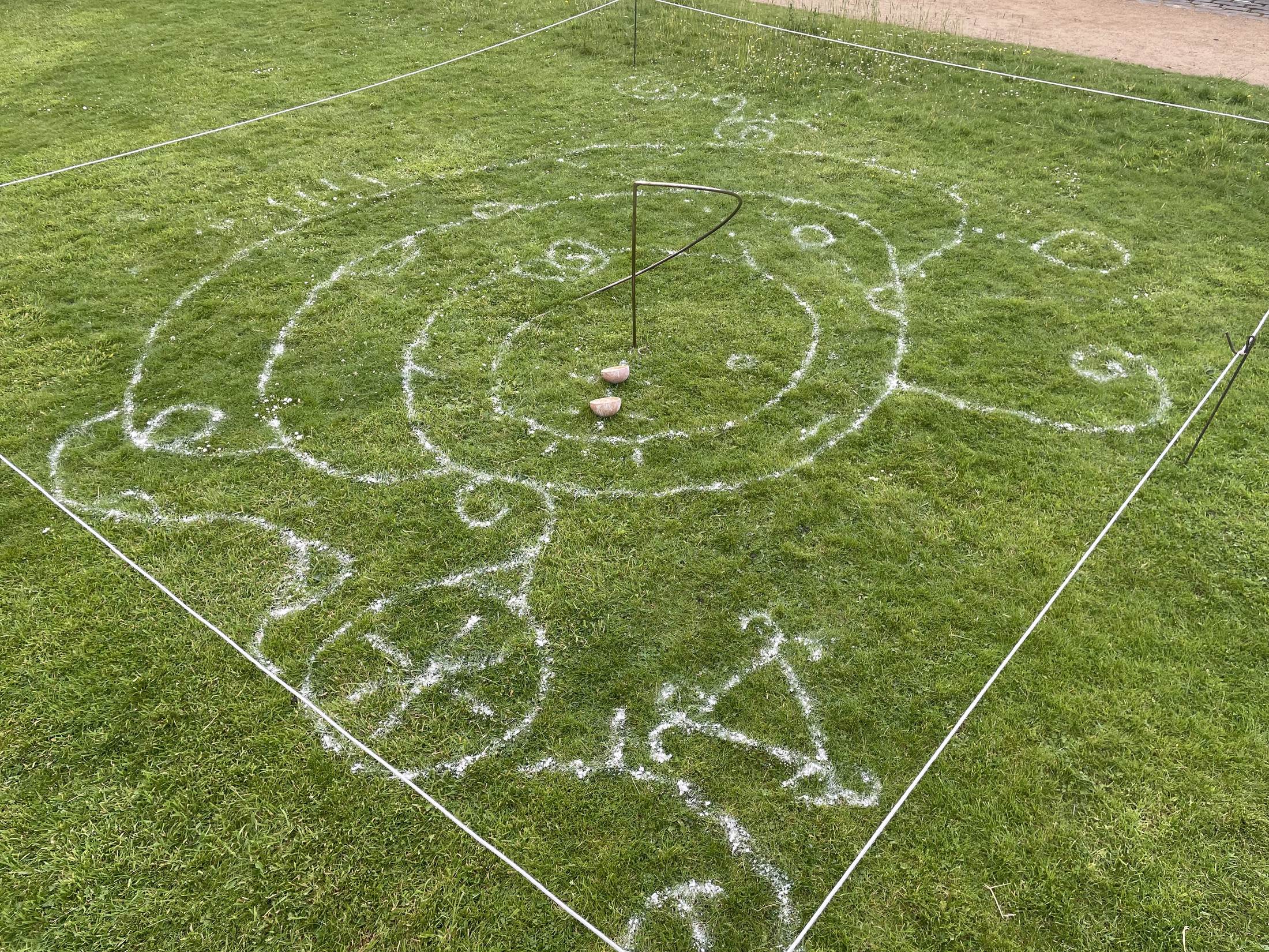 An aerial view of a large circular drawing made in white clay on grass, marked out inside a square of white cord. Curved uli lines, spirals and small circles radiate outward from a central point, where a curved bronze arc stands above two small bowls.