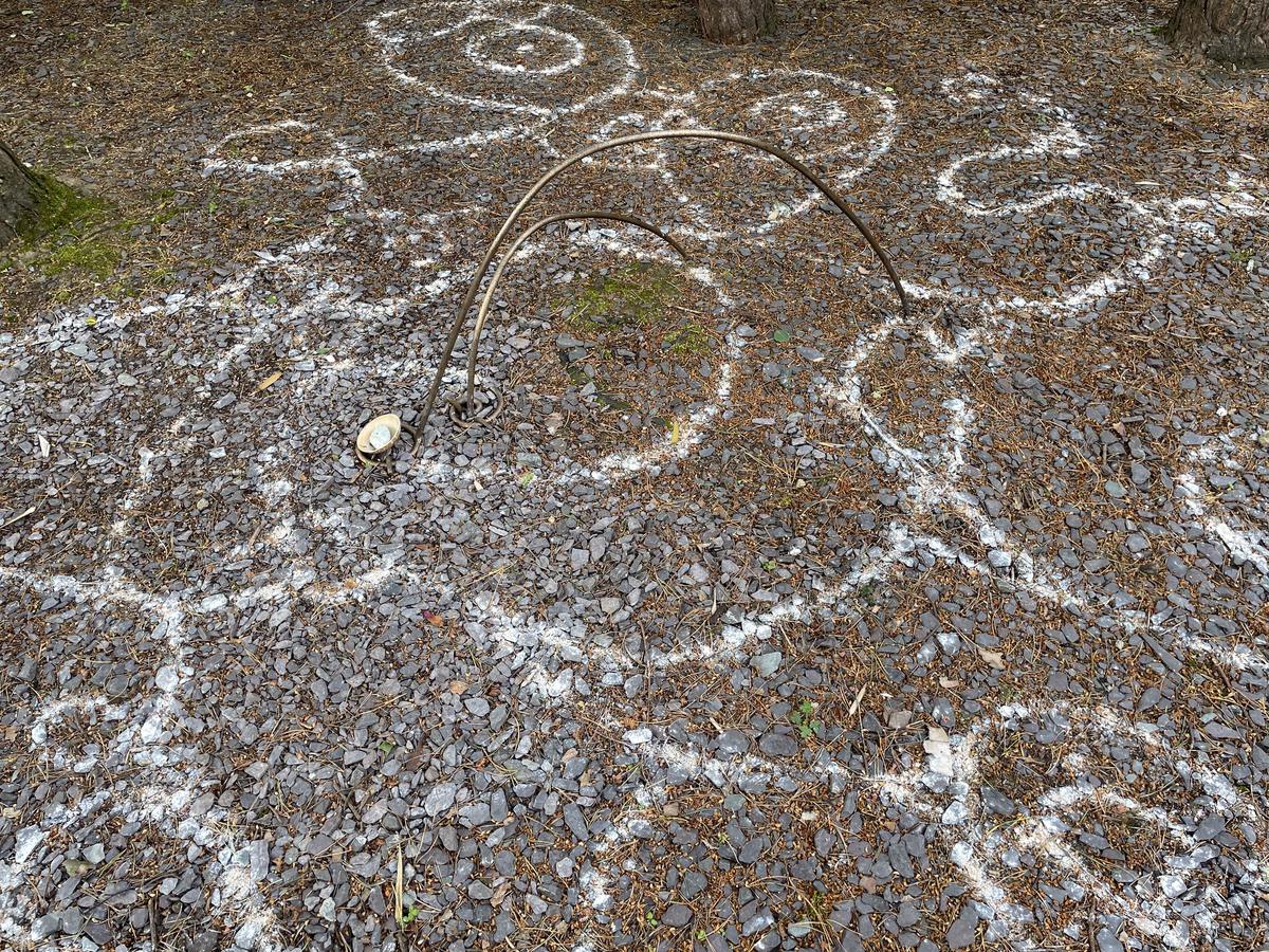 White clay lines drawn across a bed of grey slate chippings beneath trees, curving around a double bronze arc and a small pale bowl.