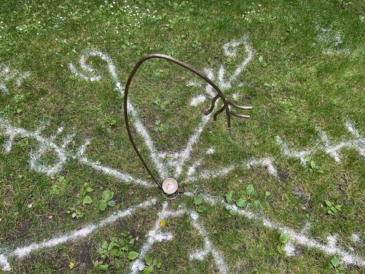 A curved bronze arc set into the grass above a small pale bowl, with white clay lines radiating outward around it.