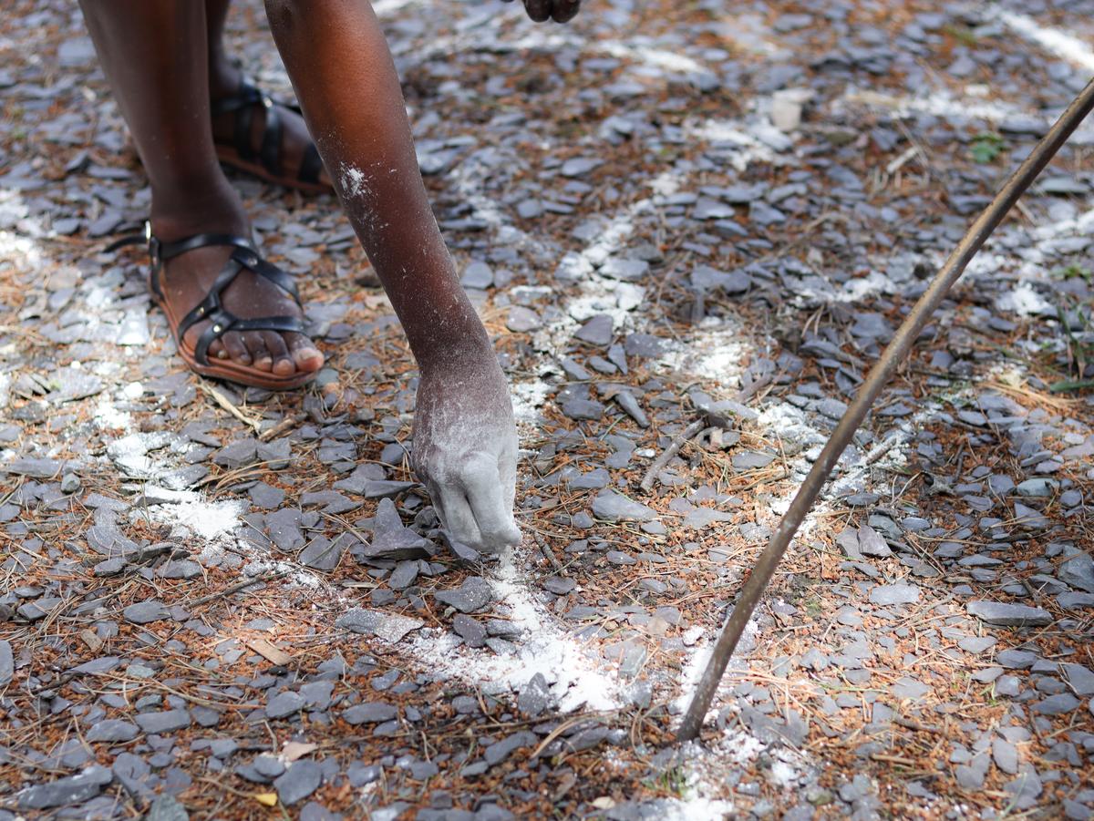 A person in sandals crouching over slate chippings, one hand white with clay, drawing a line into the ground.