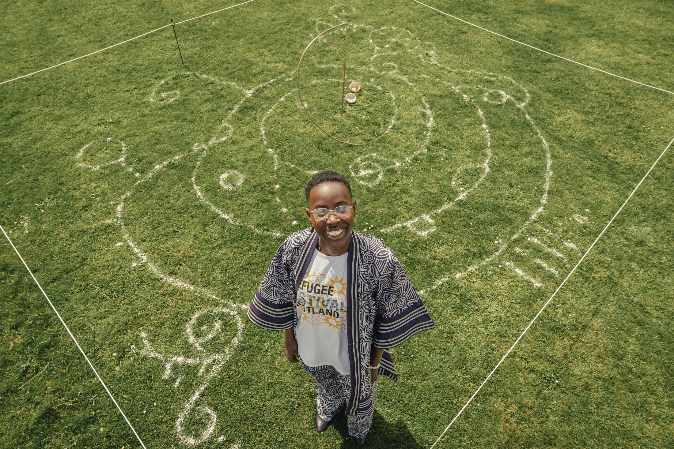 An aerial view of a circular drawing in white clay on grass, with the artist standing at its centre looking up at the camera and smiling.
