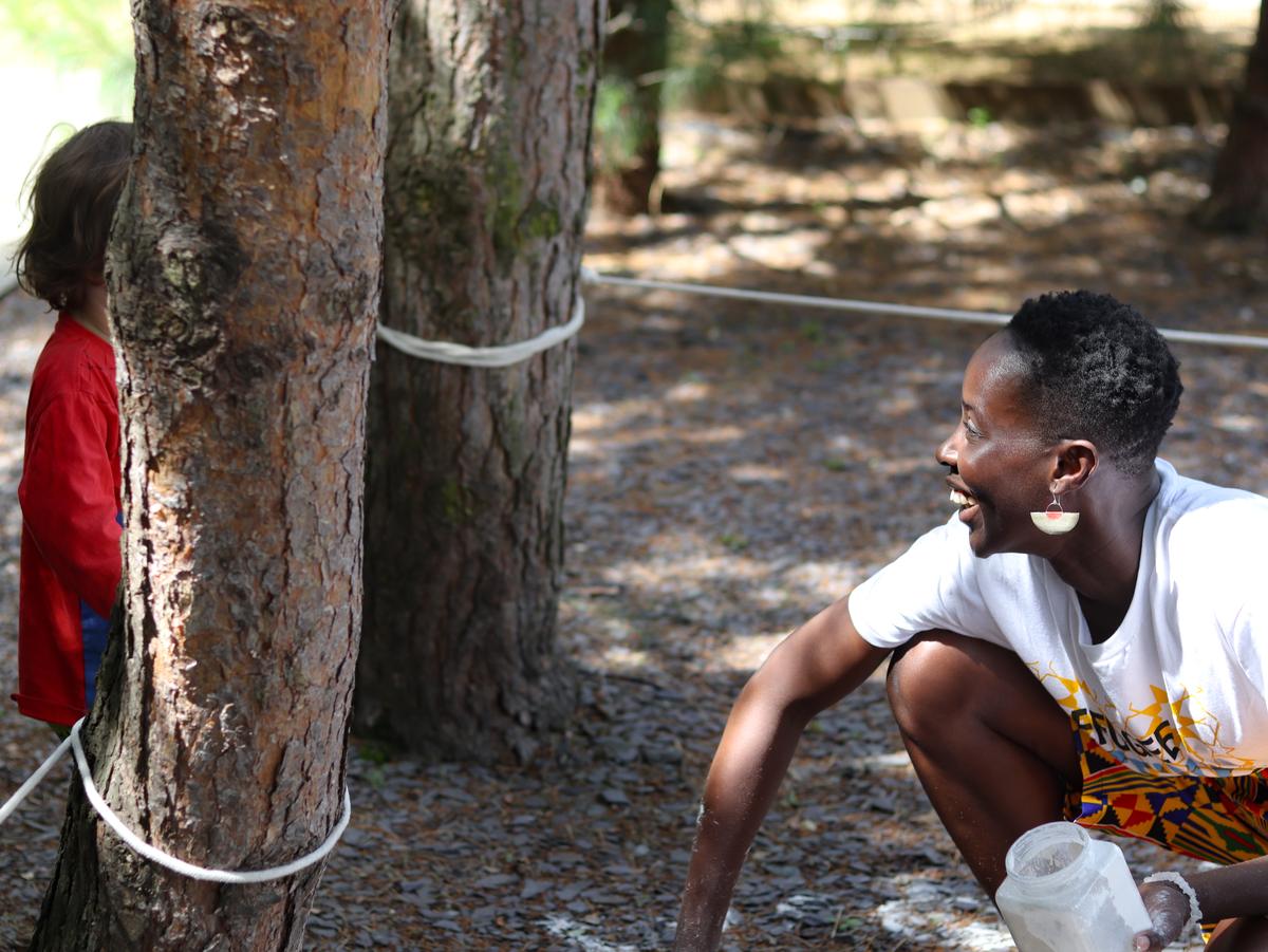 A woman in a Refugee Festival Scotland t-shirt crouching beside a pine tree, laughing, holding a jar of white clay.
