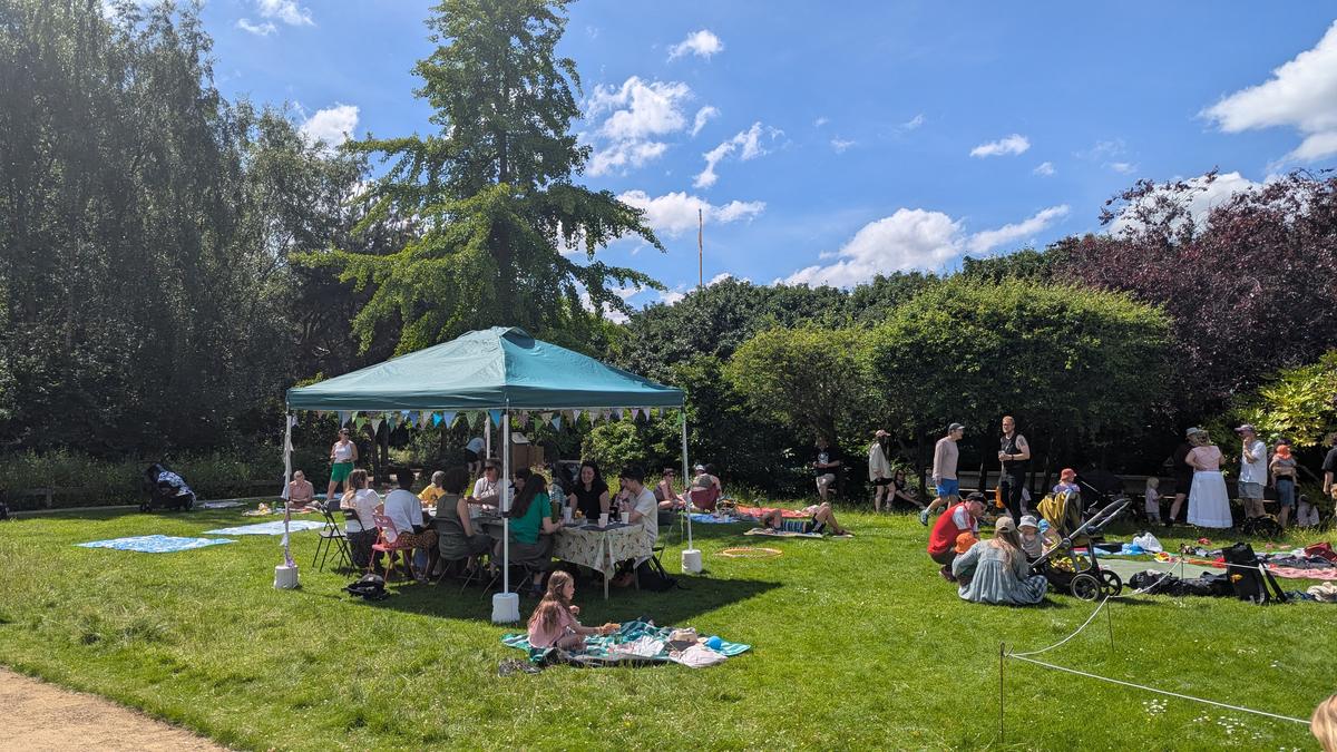 People gathered on the grass on picnic blankets around a teal gazebo with bunting, under a blue sky.
