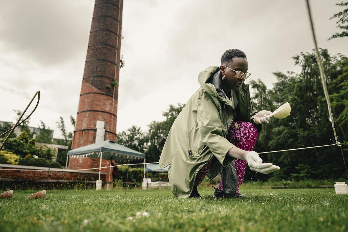 The artist crouching on the grass in a green raincoat, holding a shell of white clay and drawing a line, a brick chimney rising behind her.