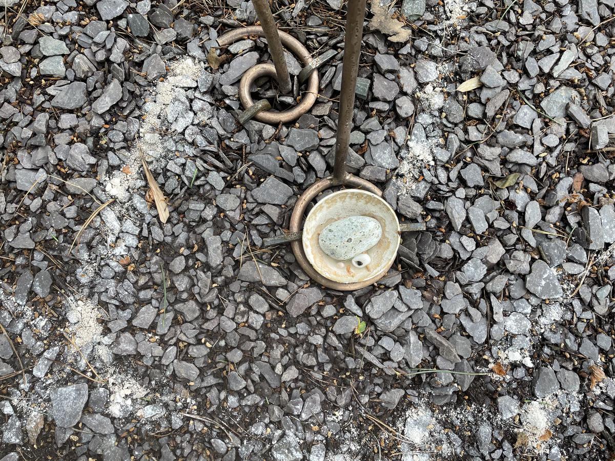 A calabash bowl set into a bronze ring on a bed of grey slate chippings, holding a pale green Iona greenstone pebble and a small cowrie shell. Bronze rods and a ring rest on the stones around it.