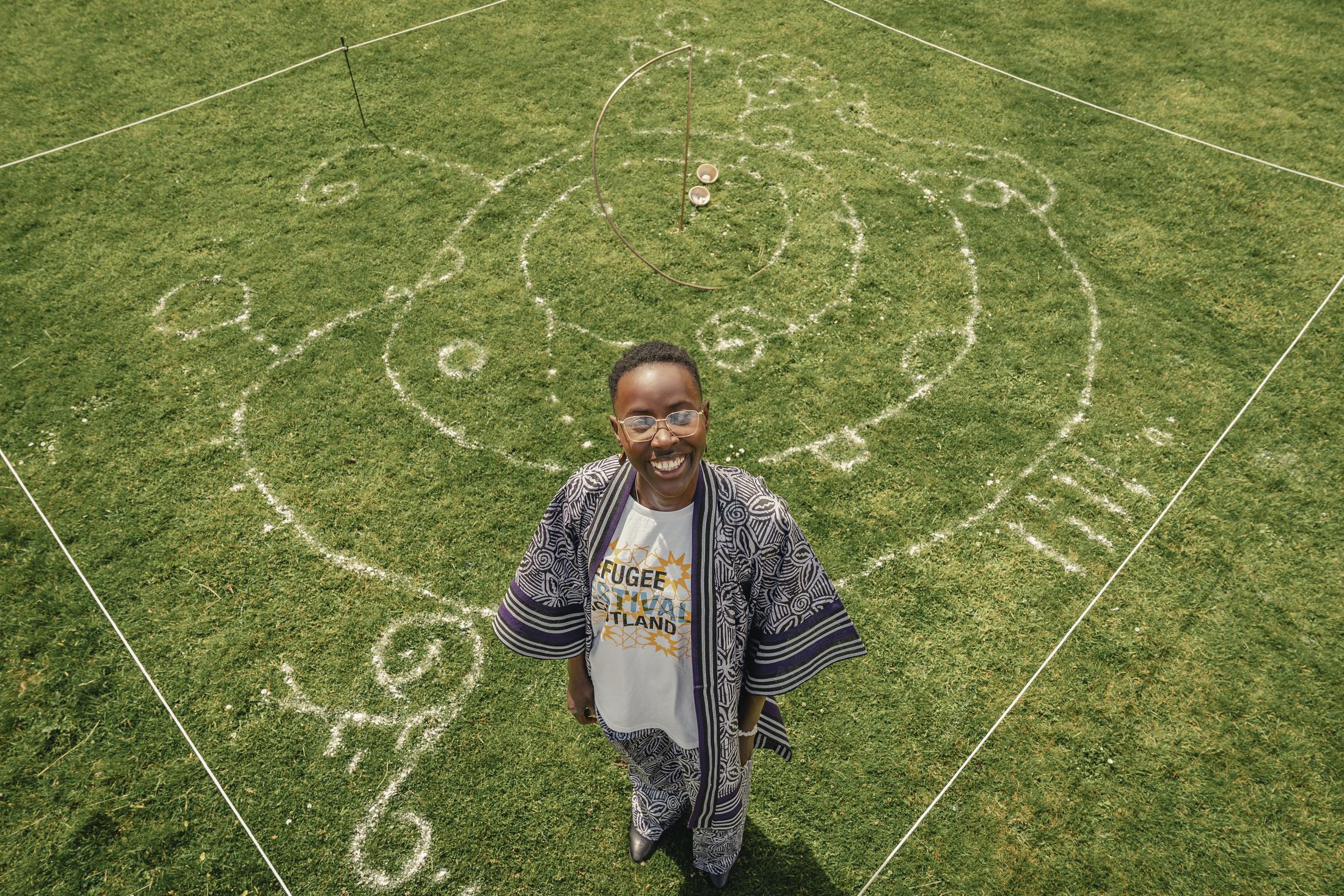 Grace Browne photographed from above, standing at the centre of a large circular drawing made in white clay on grass. Curved uli lines, spirals and small circles radiate around her, marked out inside a square of white cord. She wears a patterned indigo robe over a Refugee Festival Scotland t-shirt, and is looking up at the camera, smiling.