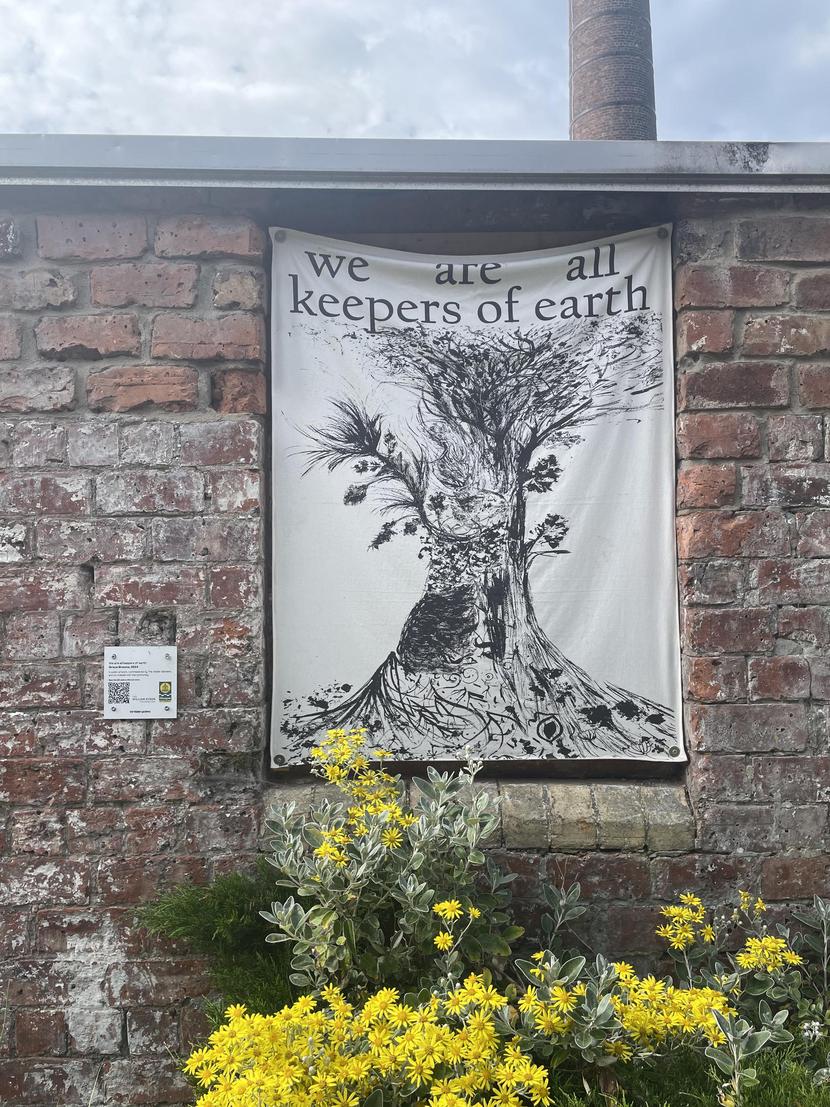 A large fabric banner hung on a weathered brick wall in the gardens. Printed on it in black is the words 'we are all keepers of earth' above a great tree with spreading roots and branches, drawn in fine ink marks. Yellow flowers grow up in front of the wall, and a small interpretation plaque with a QR code is fixed to the brick beside it.