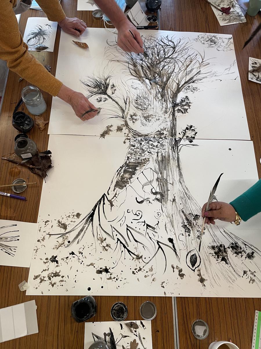 Several pairs of hands working together on a large sheet of paper, drawing a tree in black ink with brushes and foraged twigs.