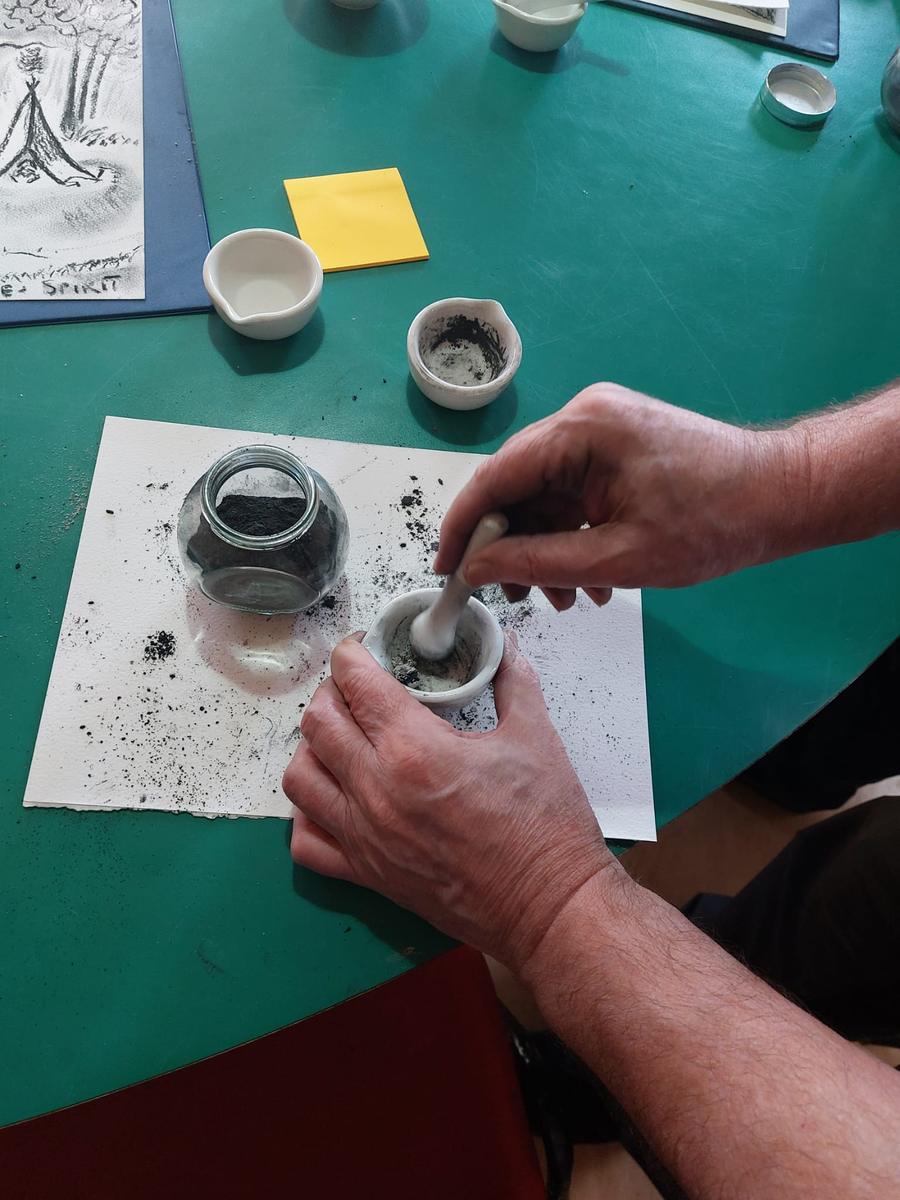 Hands grinding ash into pigment in a small mortar on a green table, beside jars and a sheet of paper.