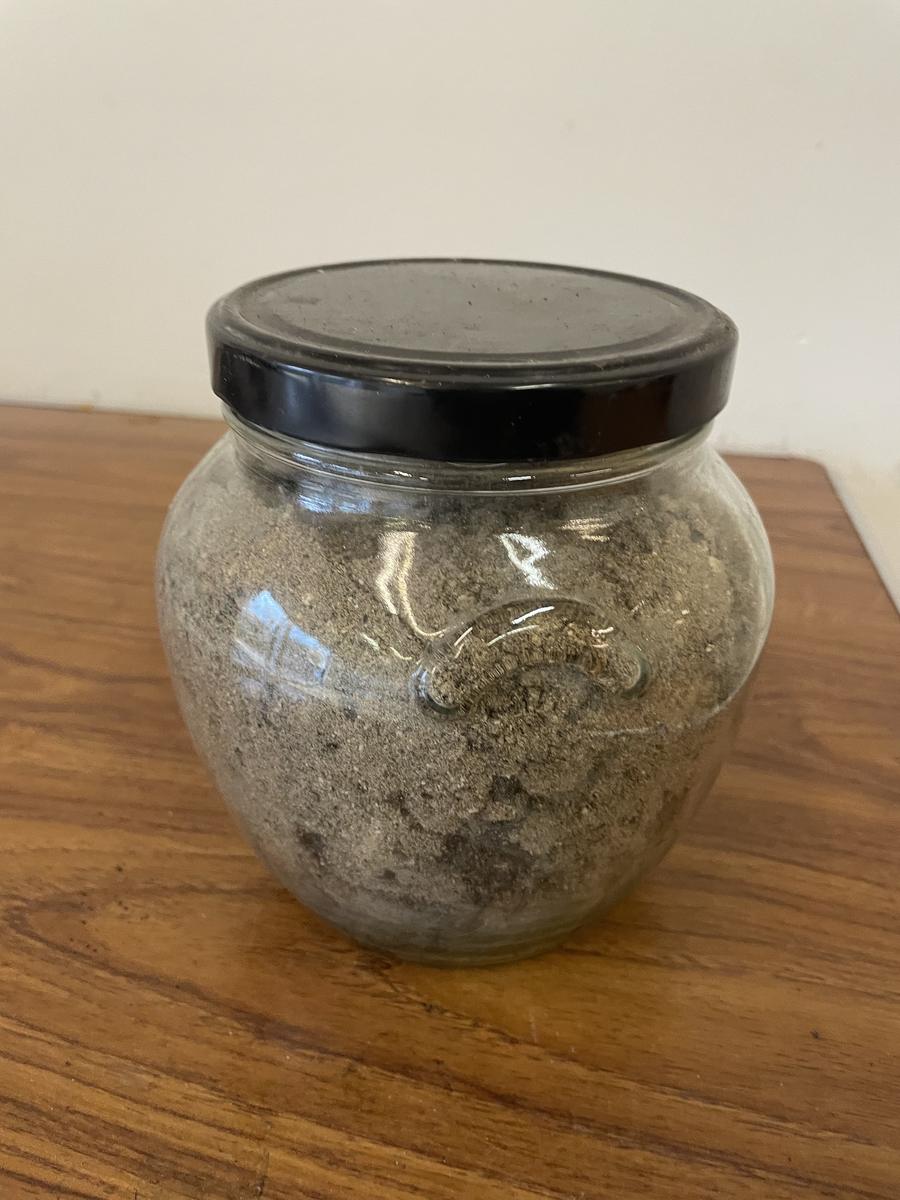 A glass jar with a black lid, filled with pale grey ash, standing on a wooden table.