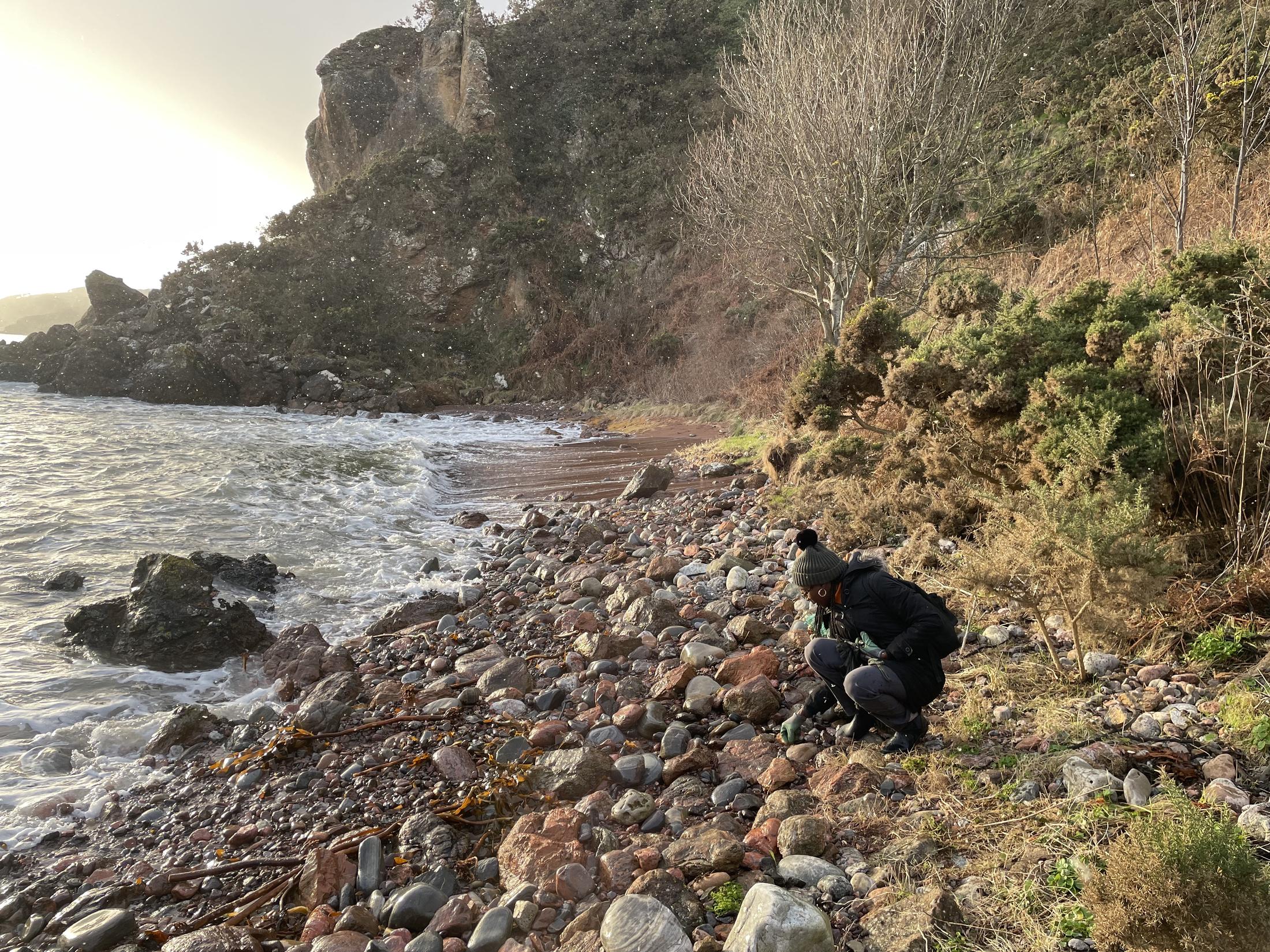 A rocky shoreline beneath a cliff at low light. A figure crouches among the boulders, gathering material from the ground.