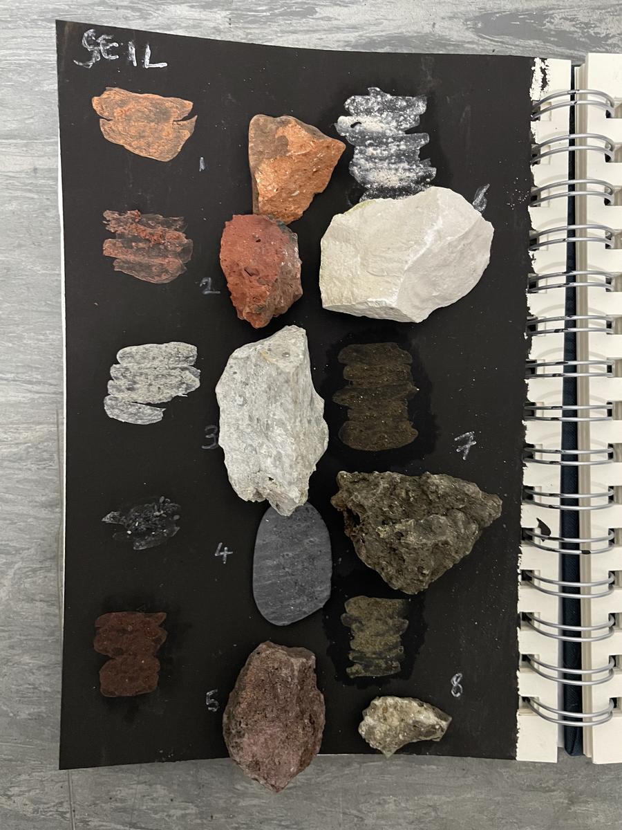 A spiral-bound sketchbook page with stones in reds, whites, greys and browns laid out in rows against black paper, annotated by hand.