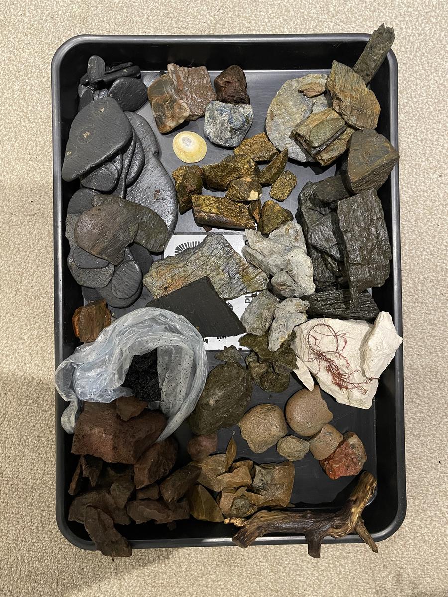 A black tray holding dozens of gathered stones, pebbles and a folded plastic bag of white clay.
