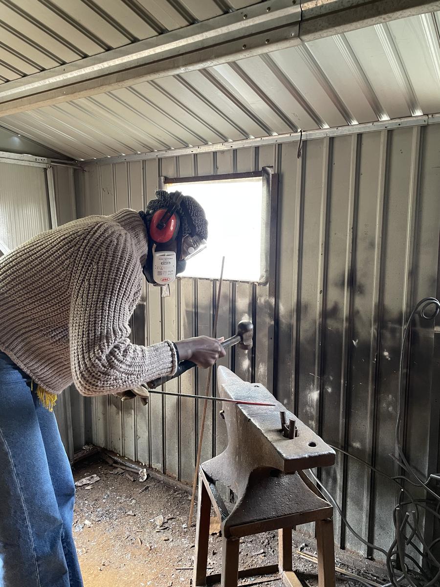 A person in a welding mask working a length of hot metal on an anvil inside a corrugated metal workshop.