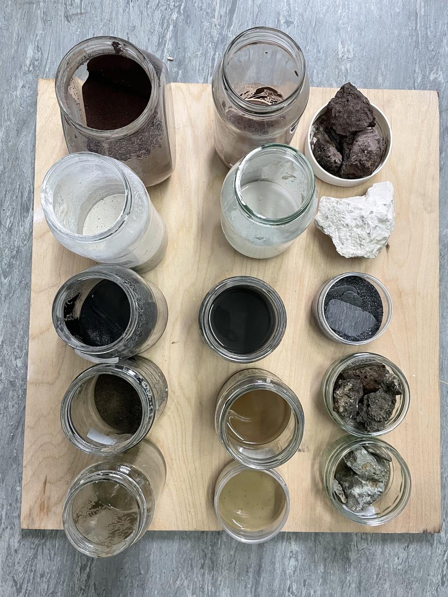 A plywood board holding twelve jars of pigment in greys, browns, ochres and black, with rock fragments alongside.