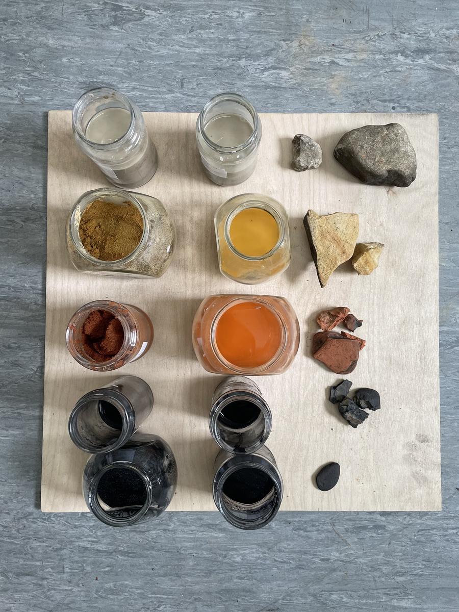 Jars of ground pigment and liquid paint in creams, golden yellow, orange and black on a plywood board, each beside its parent stone.