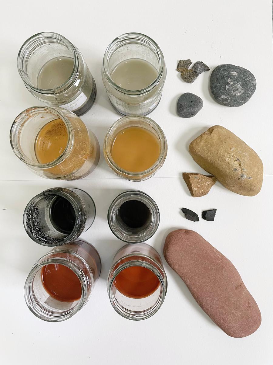 Eight jars of liquid pigment in grey, gold, black and deep red on white, with the pebbles and charcoal they came from.