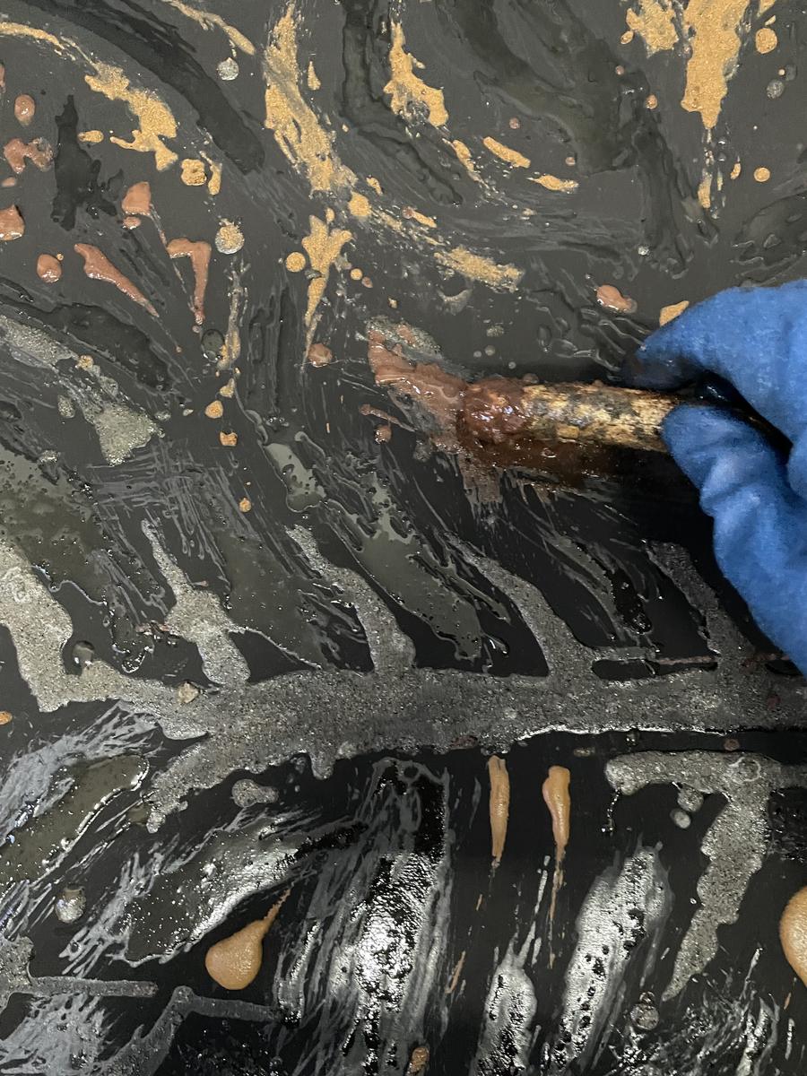 A hand in a blue glove drawing with a stick through wet black paint, revealing gold and red pigment beneath.