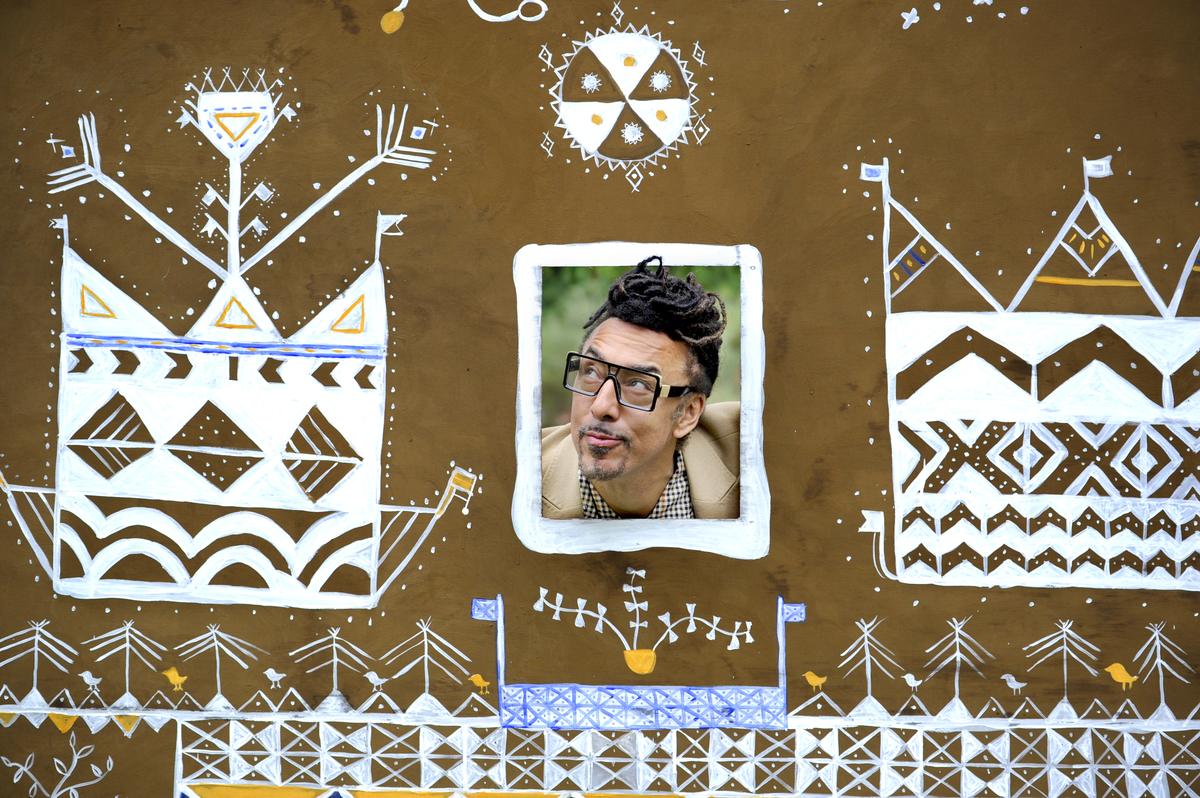 A woman in a patterned dress leaning through an opening in the brightly painted Ndebele-inspired panel, taking a photograph of herself, the other panels behind her.