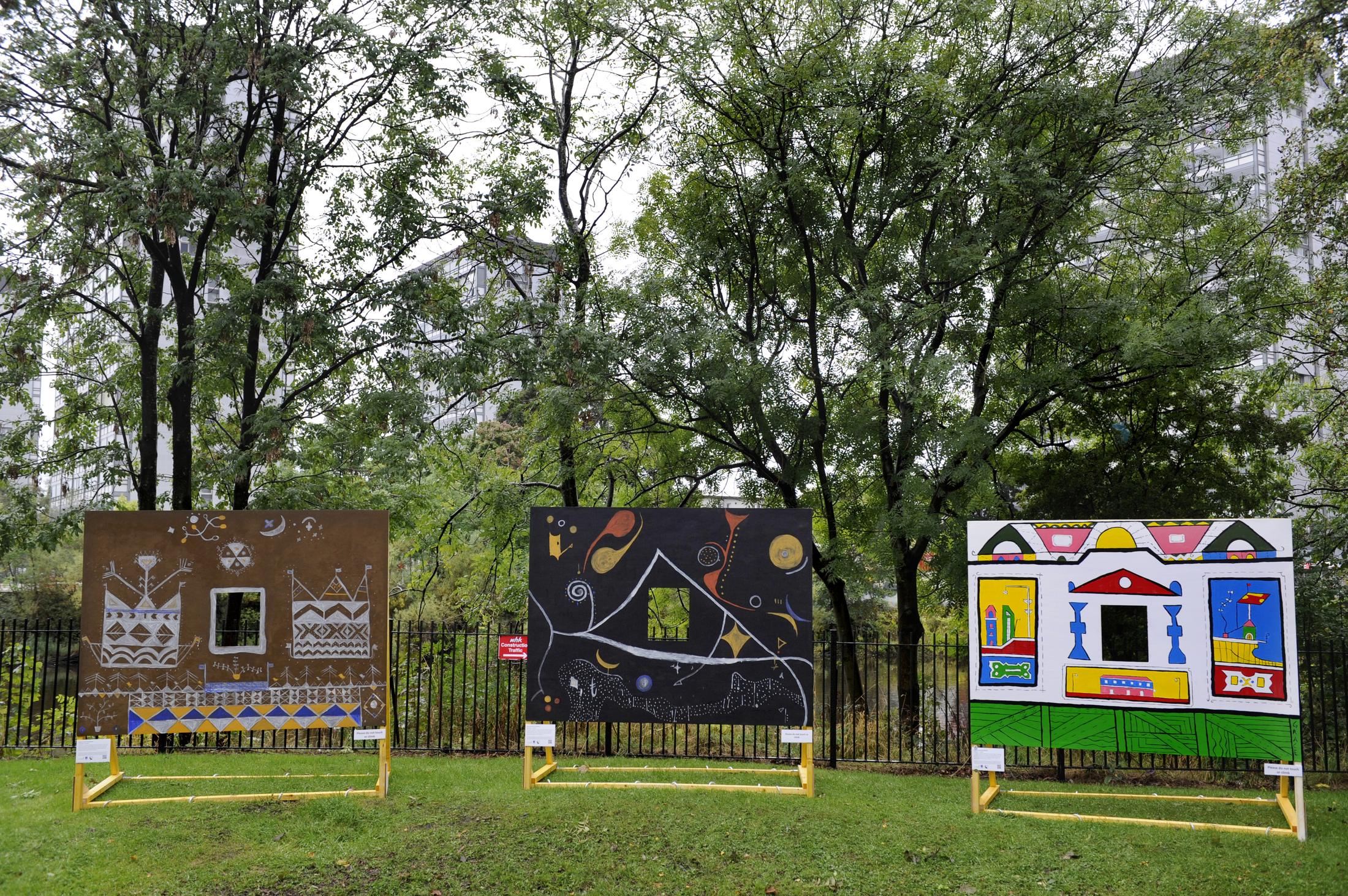 Three large painted mural panels standing on yellow frames in a park among tall trees. The first is brown with white geometric pattern, the second black with flowing figures in orange and white, the third white and green with bright architectural shapes.