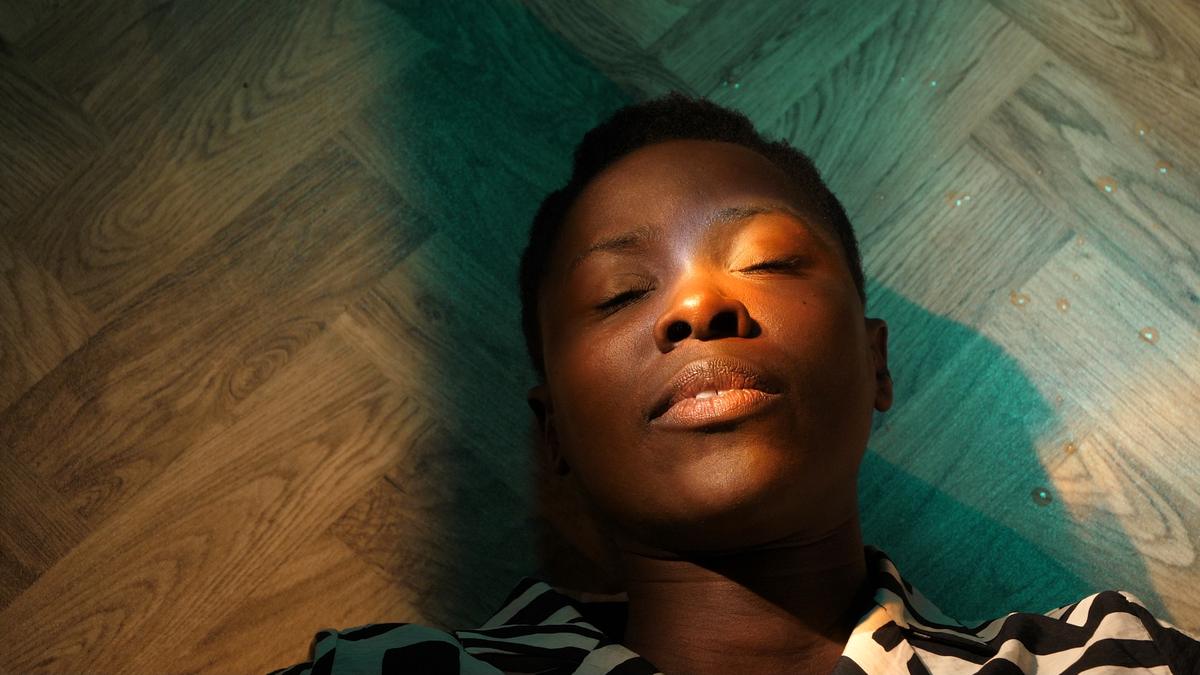 A film still: a woman lying on a parquet floor with her eyes closed, warm orange light falling across her face, a green cloth beneath her.
