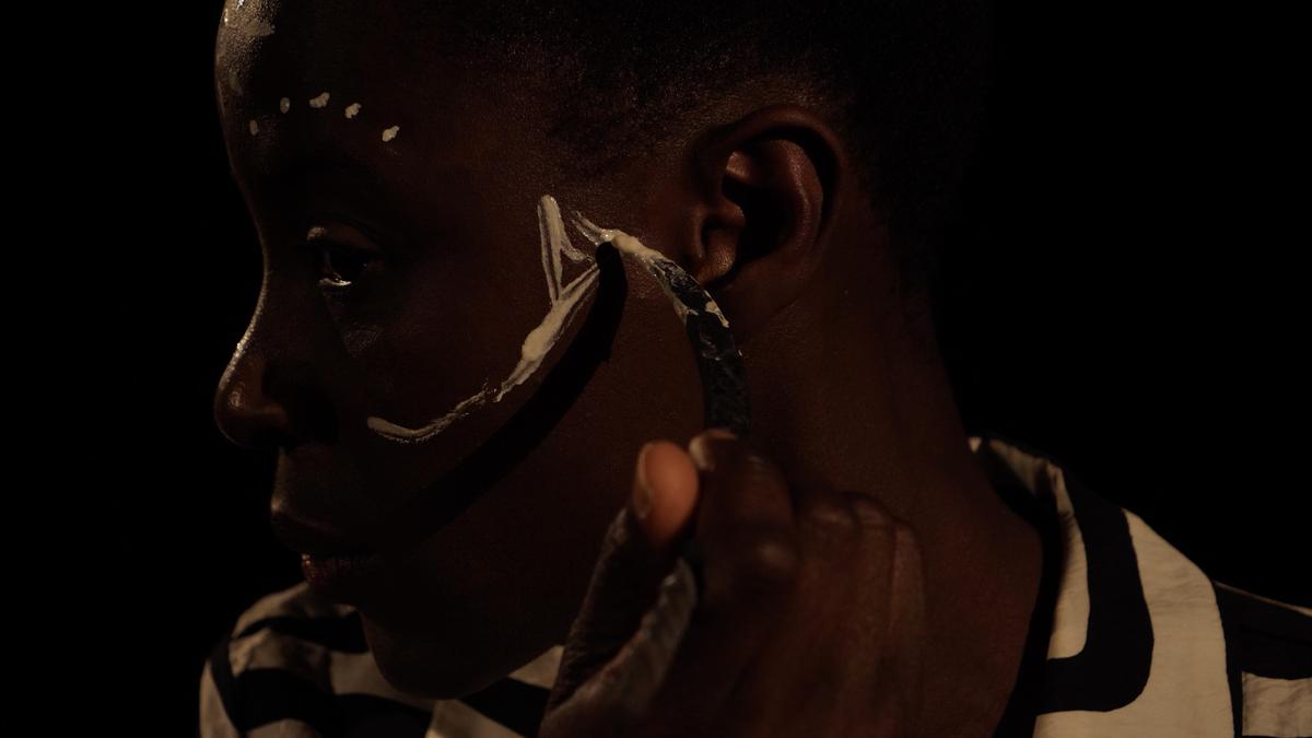 A film still in near darkness: a woman in profile, a row of small white dots marked across her temple, holding a curved shell or blade to her ear.