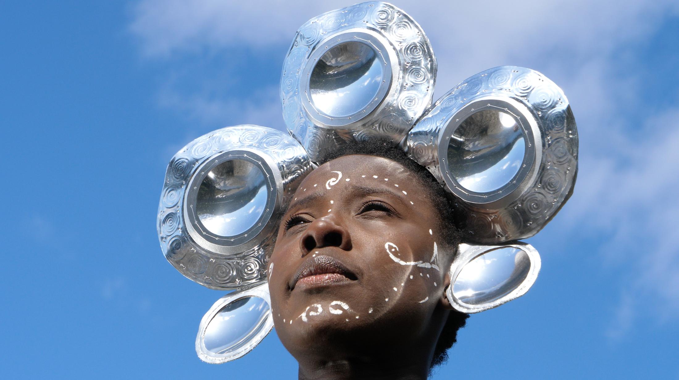 A film still: a woman photographed from below against a blue sky, wearing a headdress of large polished metal discs. Fine white uli marks curl across her cheeks and forehead.