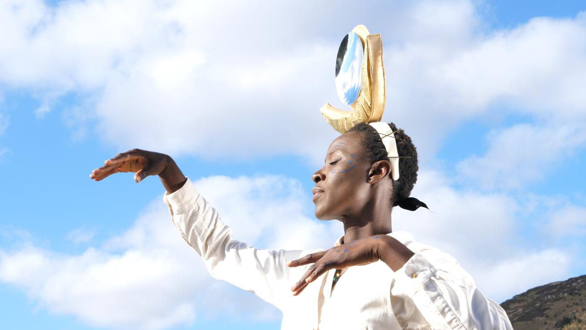 A film still: a woman in white against a bright blue sky and clouds, arms raised and curved like wings, wearing a tall gold and mirrored headpiece.