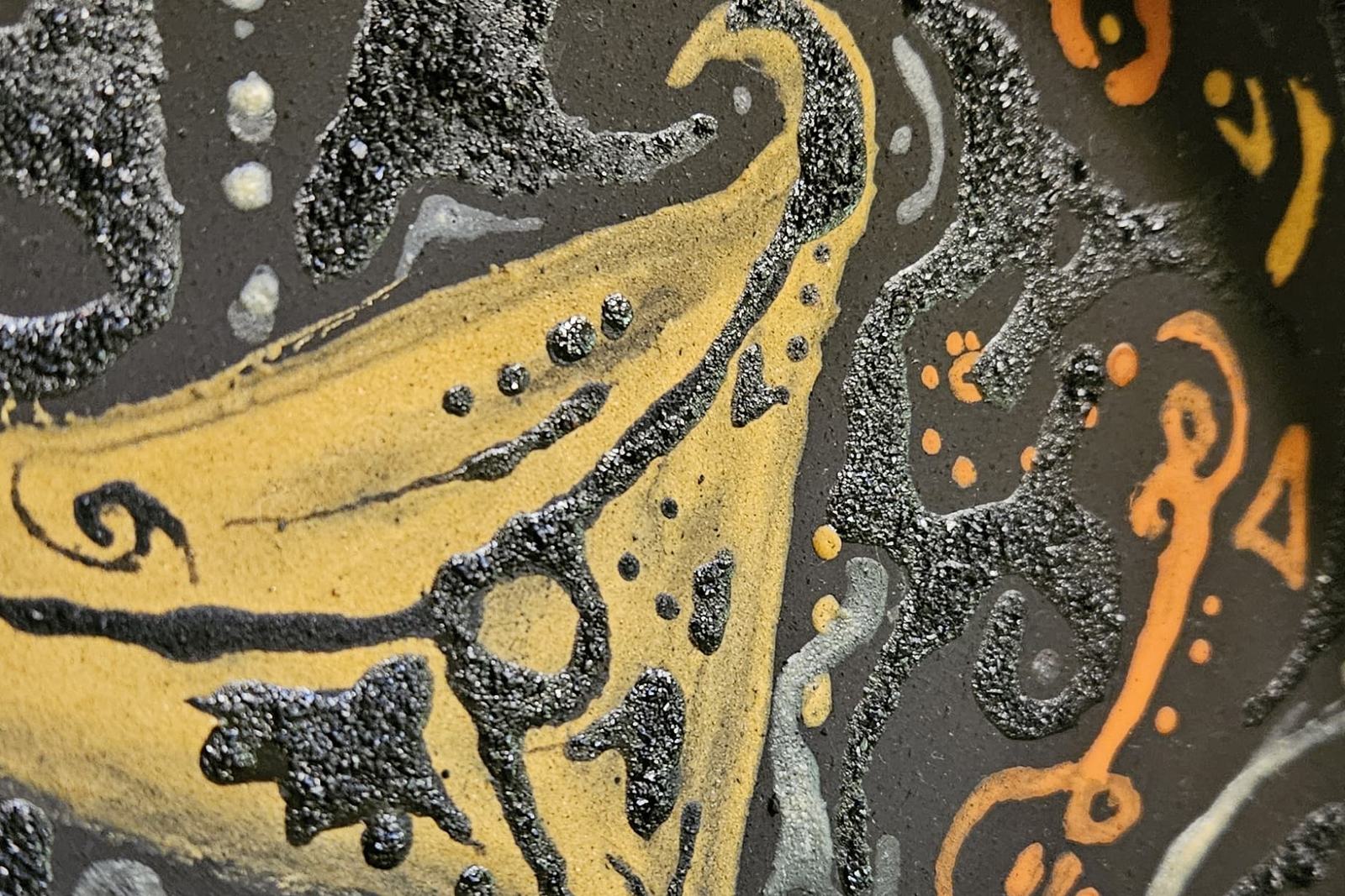 A close view of a painted wooden bowl: a curved yellow form outlined in black on a dark speckled ground, with fine white and orange marks around it.