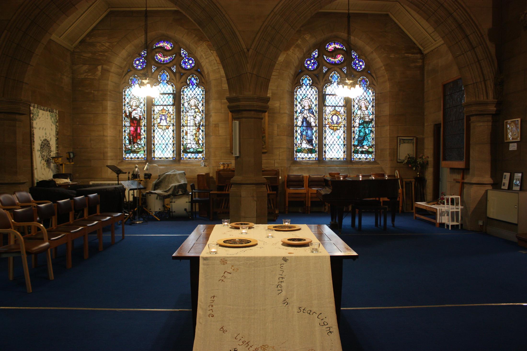 The interior of a stone church with two stained glass windows. A long table runs down the centre, covered with unbleached cotton cloth and set with painted wooden bowls.