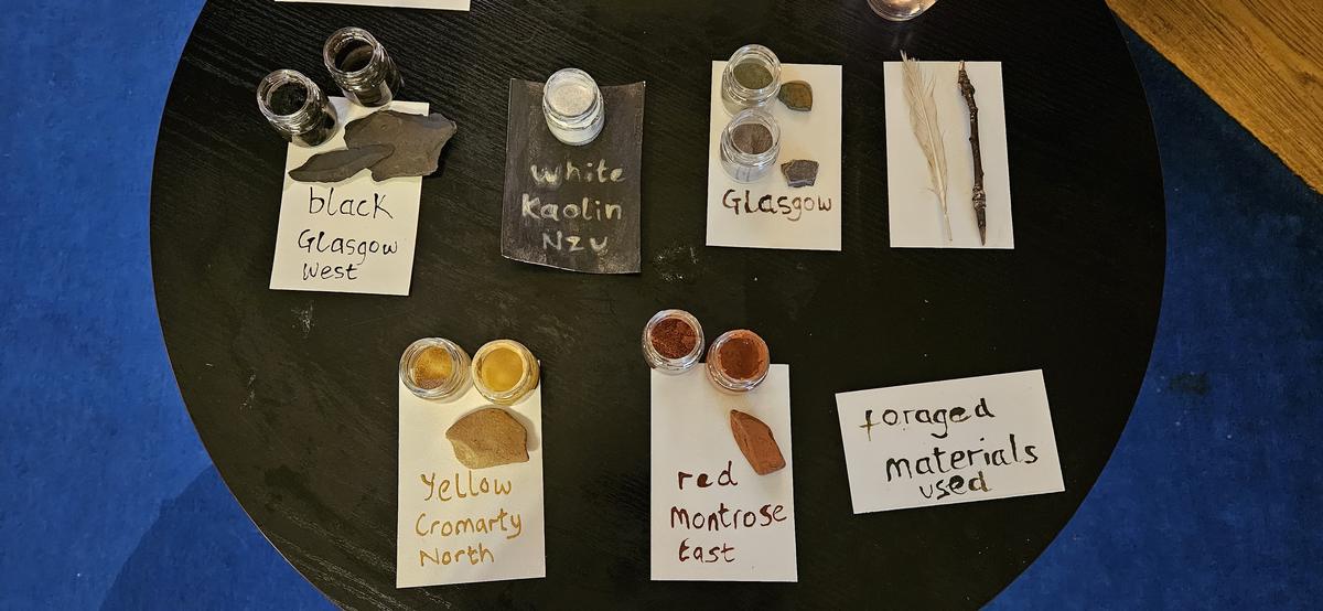 A dark round table seen from above, laid with small glass jars of ground pigment beside the rocks they came from, each on a handwritten card: black Glasgow west, white kaolin nzu, Glasgow, yellow Cromarty north, red Montrose east. A feather and a stick lie on a card marked foraged materials used.
