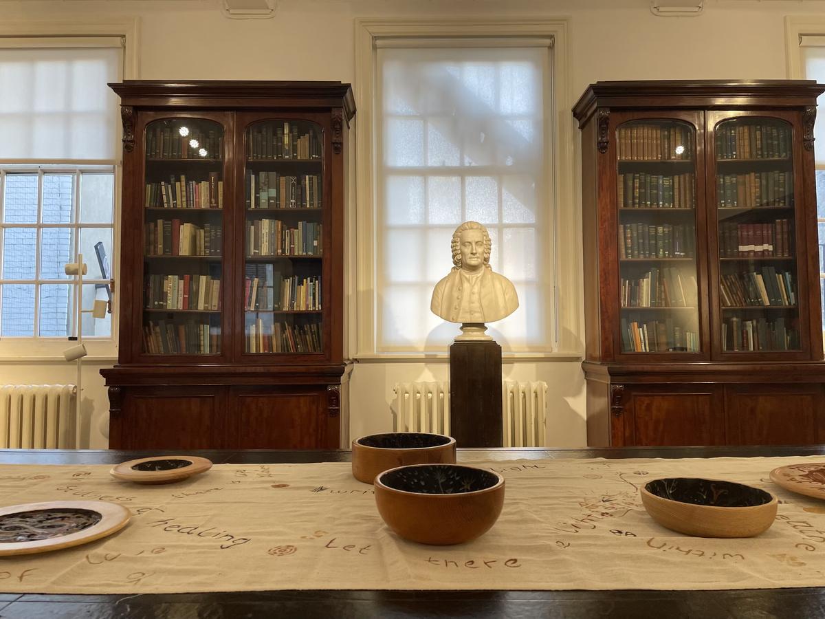 A panelled library with tall glazed bookcases and a marble bust between two windows. A long table in front holds several painted wooden bowls on a length of cloth.