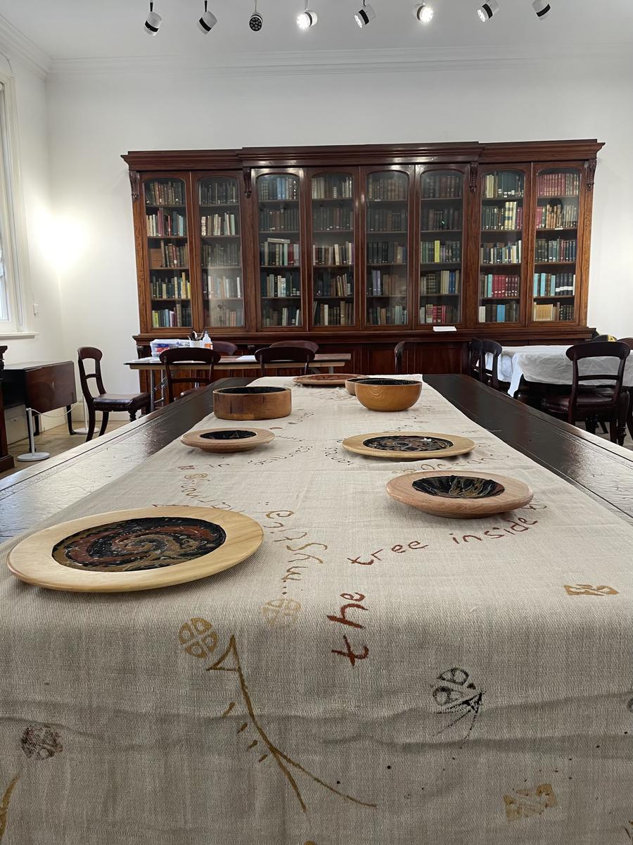 The long table seen from one end, the painted bowls set out along a cloth marked with handwritten script, the library bookcases behind.