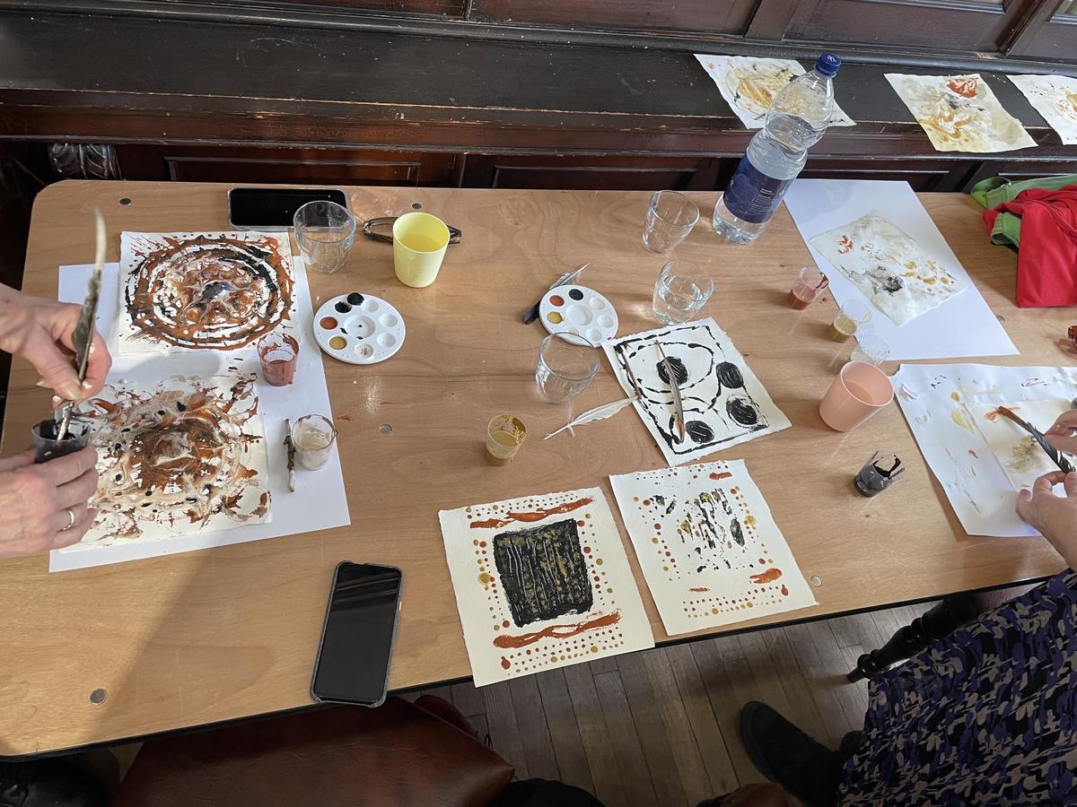 A long table in a panelled room covered with paintings in earth pigment, palettes, jars and feather quills, with several people working around it.