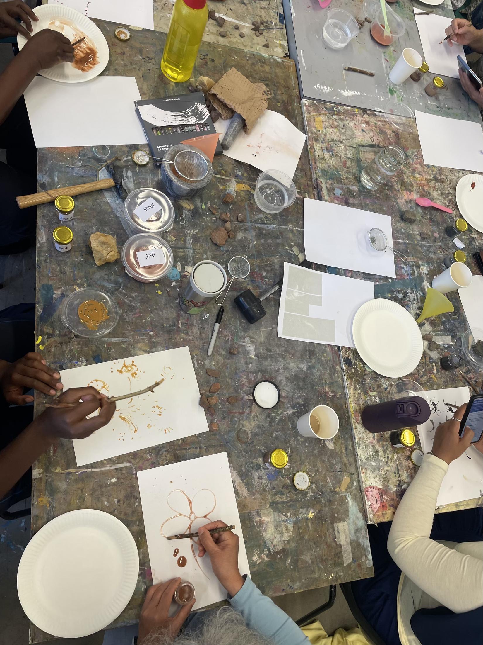A workshop table seen from above, covered in paper, jars, palettes and brushes. Several pairs of hands are painting with earth pigments in browns and ochres.
