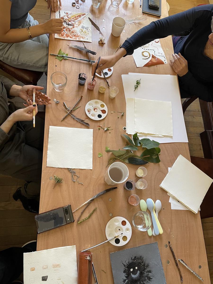 A long wooden table seen from above during a workshop, with palettes, brushes, foraged materials and paintings in progress spread across it, and several people working around the edges.