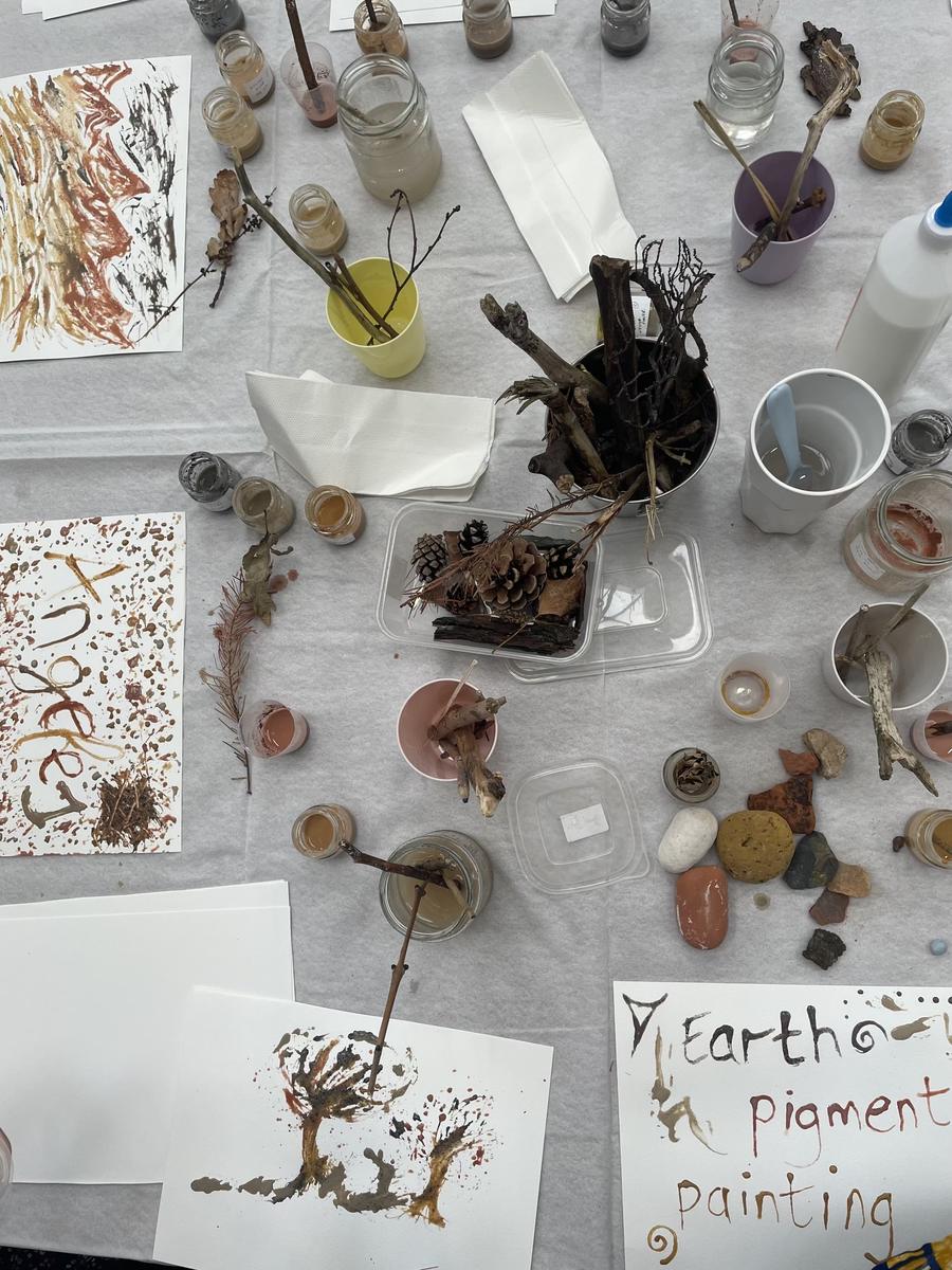 Paintings drying on a table beside jars, brushes and a handwritten sign reading 'earth pigment painting'.