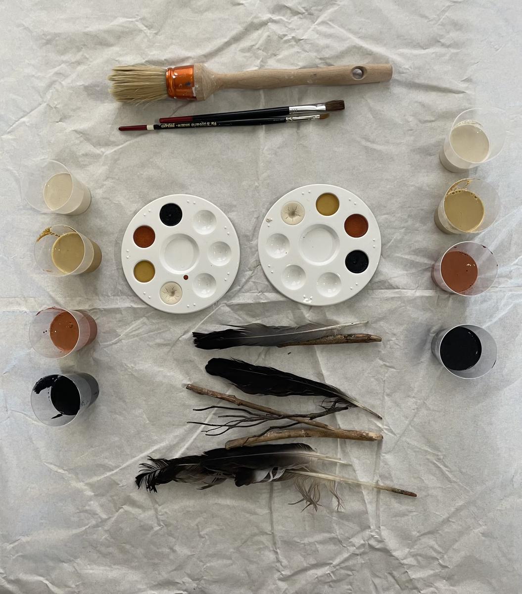 Two white ceramic palettes surrounded by small pots of earth pigment in ochres, reds, browns and black, with brushes and feathers laid out below.