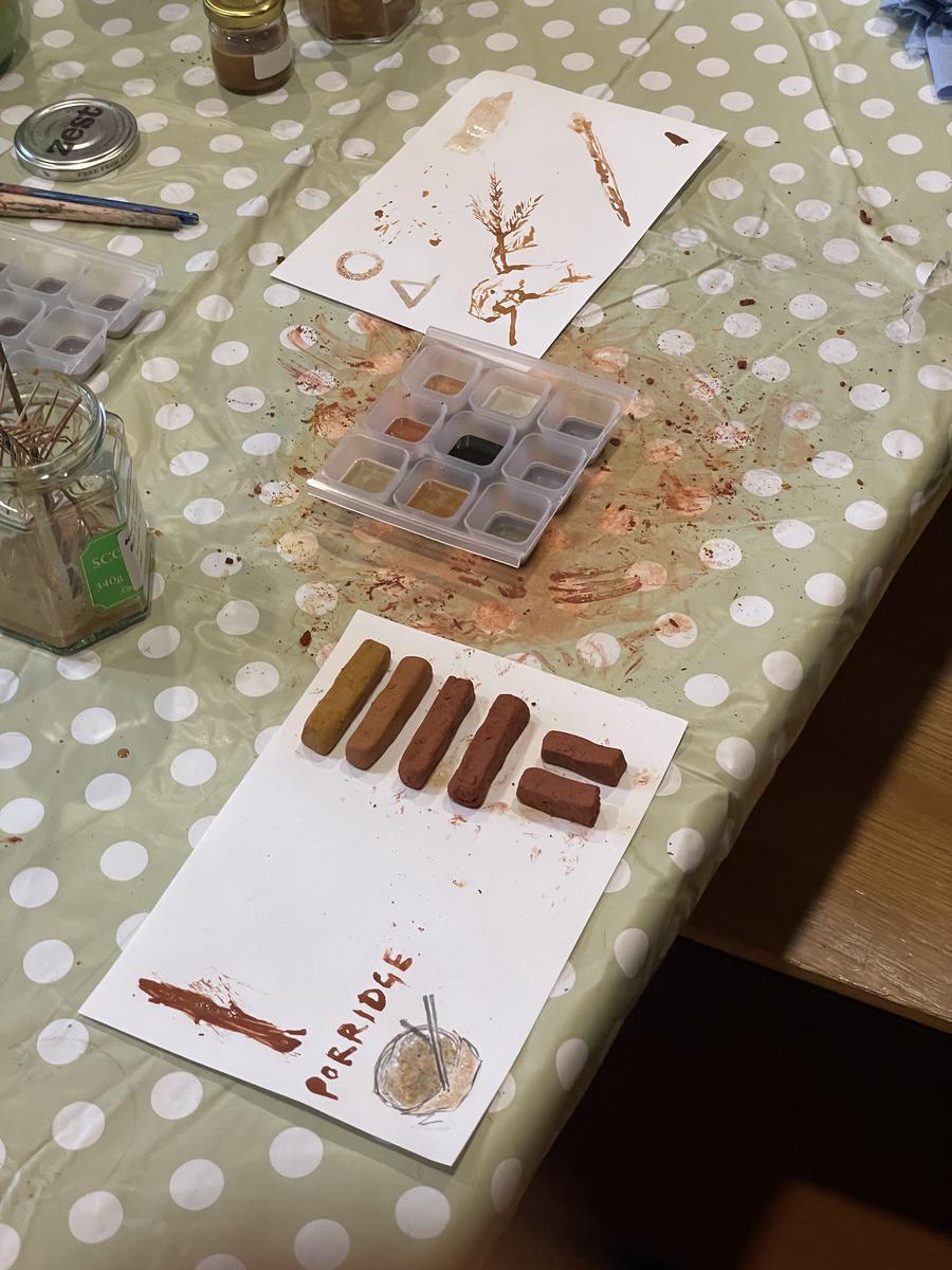 Sticks of hand-rolled earth pastel in ochre and terracotta drying on a sheet of paper, beside a painted colour swatch labelled 'porridge'.