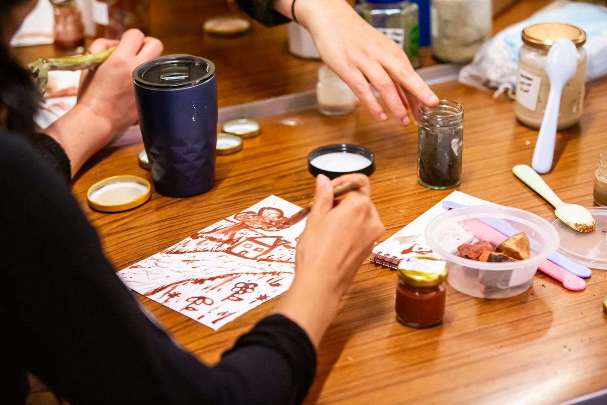 Hands at a workshop table stirring terracotta pigment in a small glass jar with a twig, surrounded by labelled jars of pigment, a water bottle and a sheet of test marks.