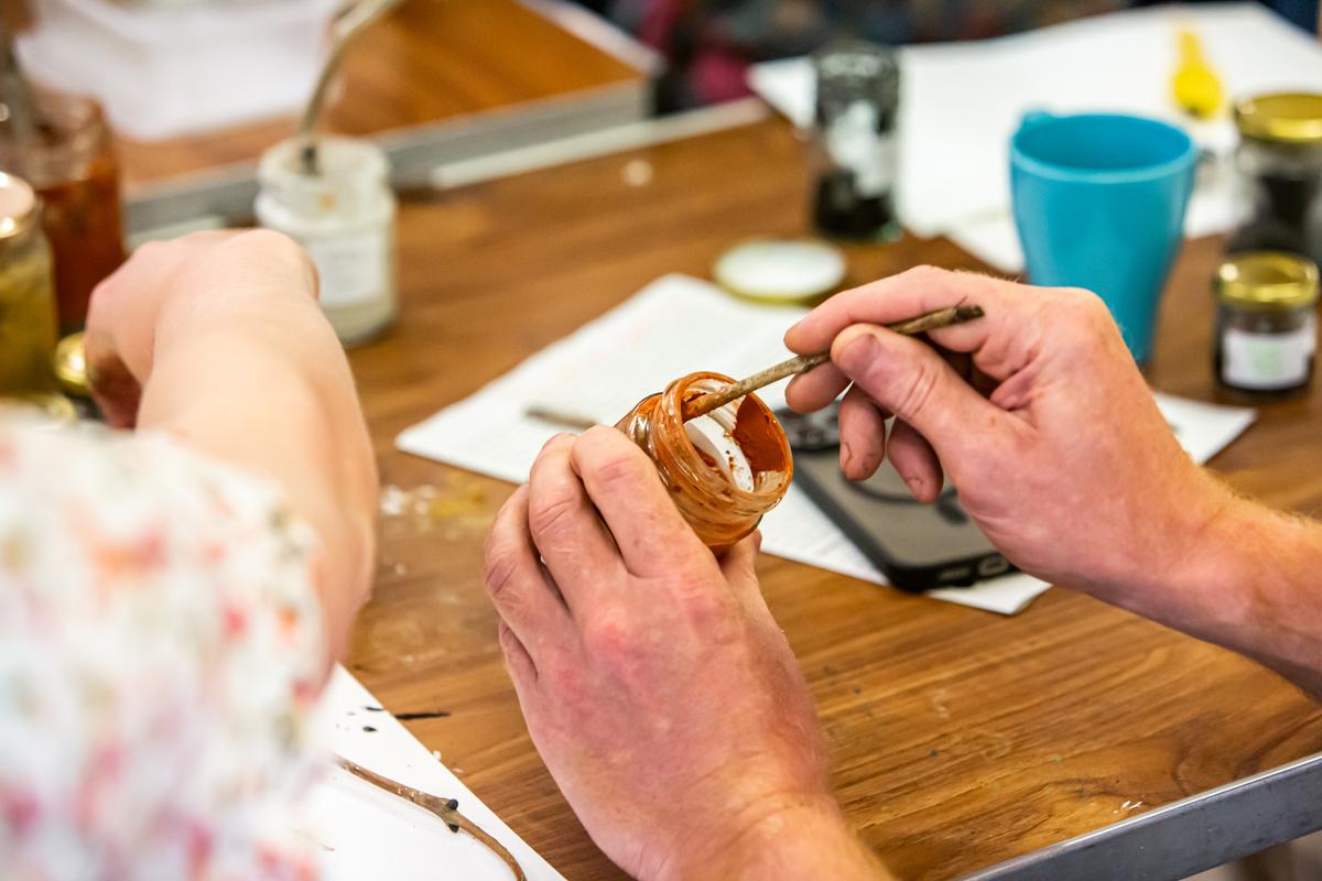 A person painting a house and garden in red earth pigment on white paper, using a twig as a brush, with jars of pigment and a tub of rock fragments beside them.