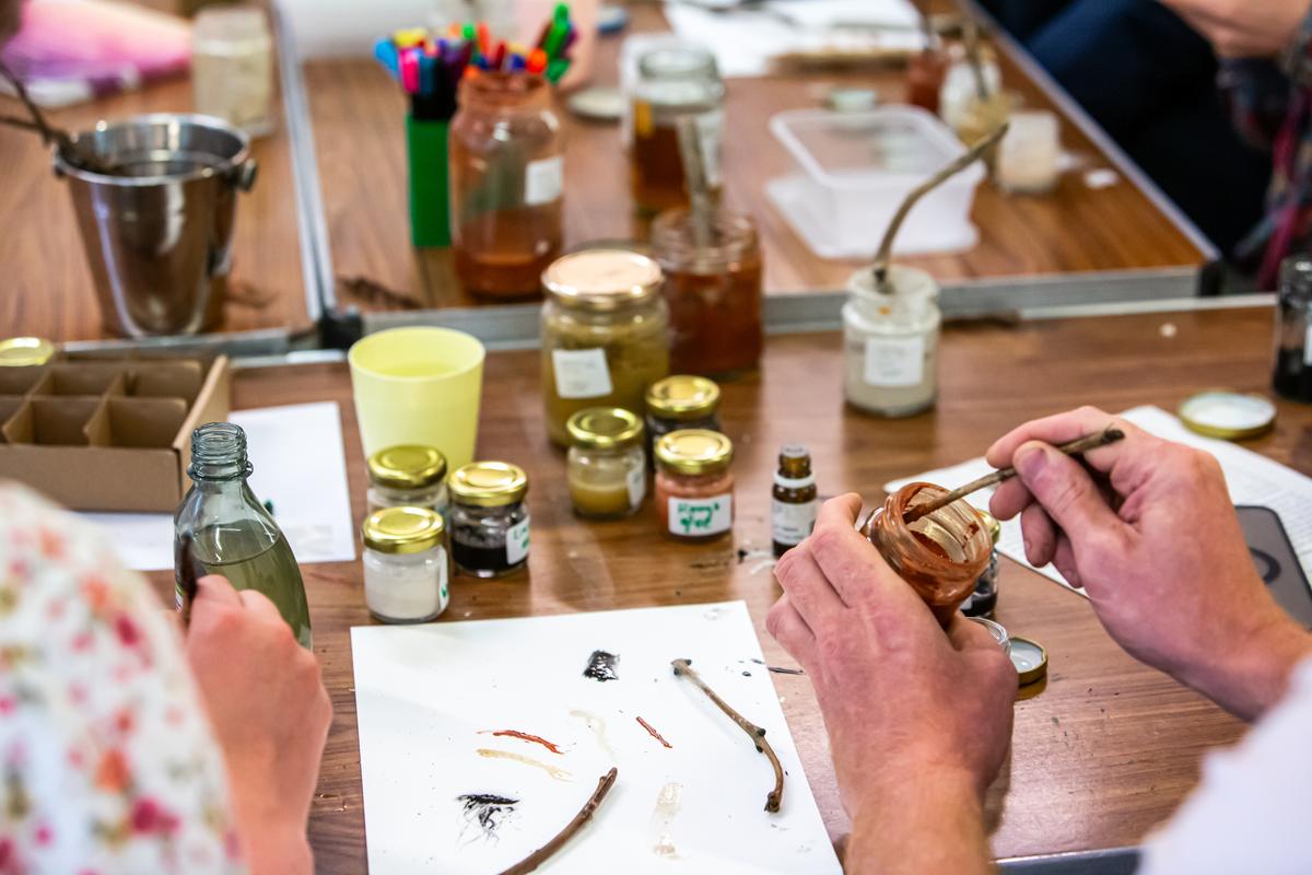 Close view of two hands holding a jar of terracotta pigment and drawing a twig through it to load the tip, at a workshop table scattered with jars and paper.