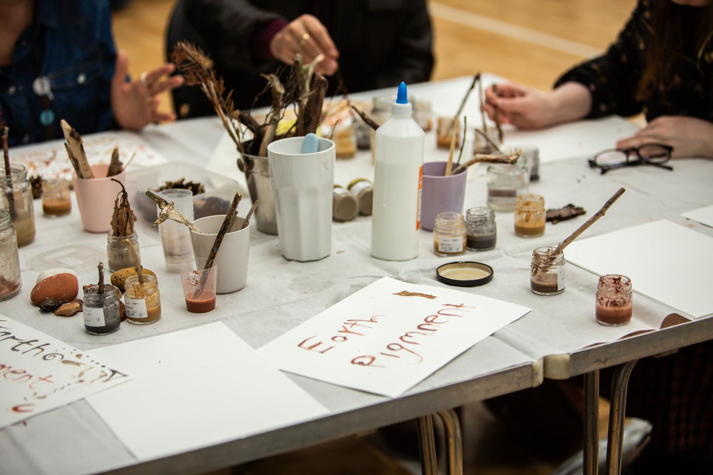 A workshop table covered in white cloth, laid with jars of earth pigment, twigs and sticks standing in pots, and sheets of paper. One sheet is painted with the words 'Earth Pigment' in red ochre. People are working around the table.