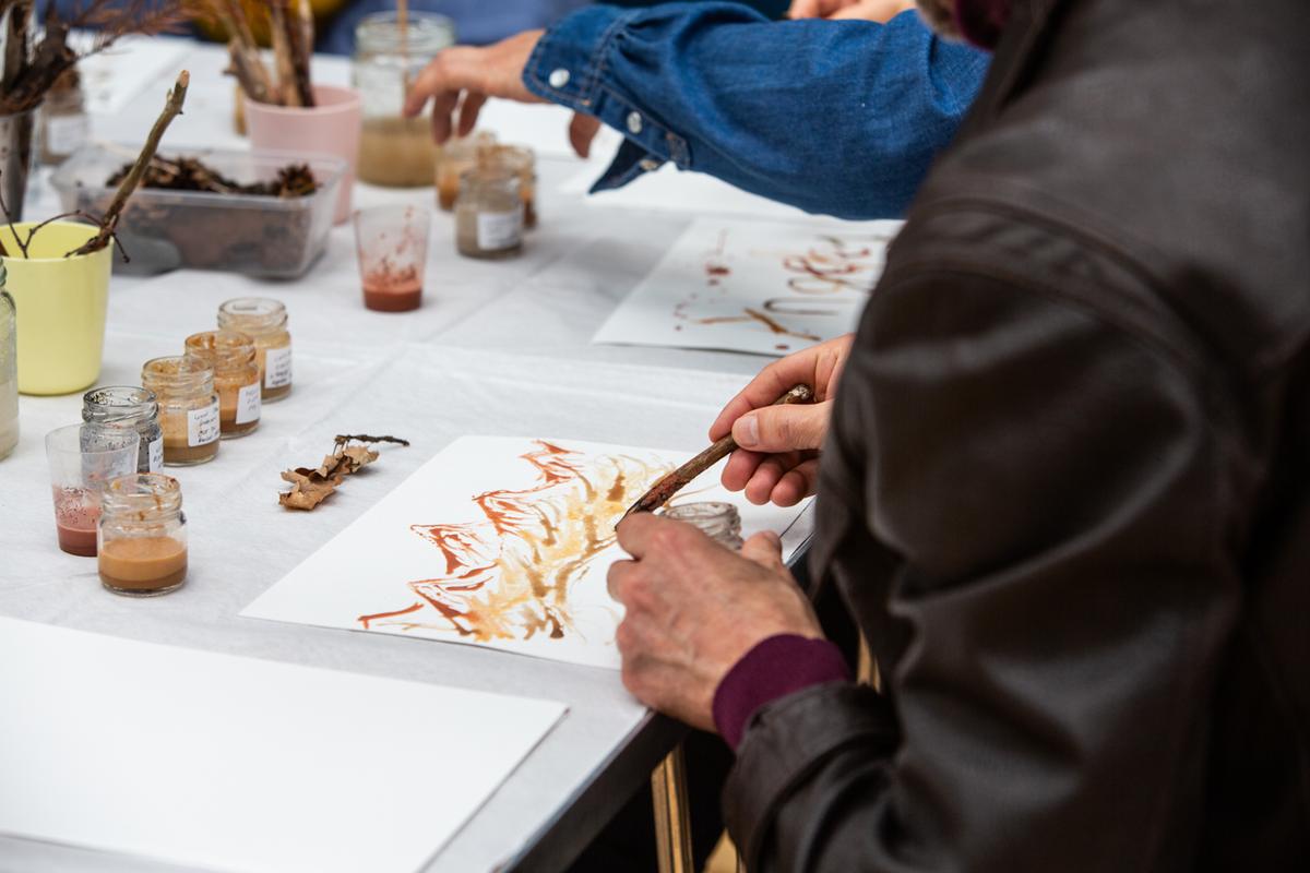 A person drawing in orange and ochre earth pigment on white paper with a twig, at a workshop table set with labelled jars of pigment and foraged materials.